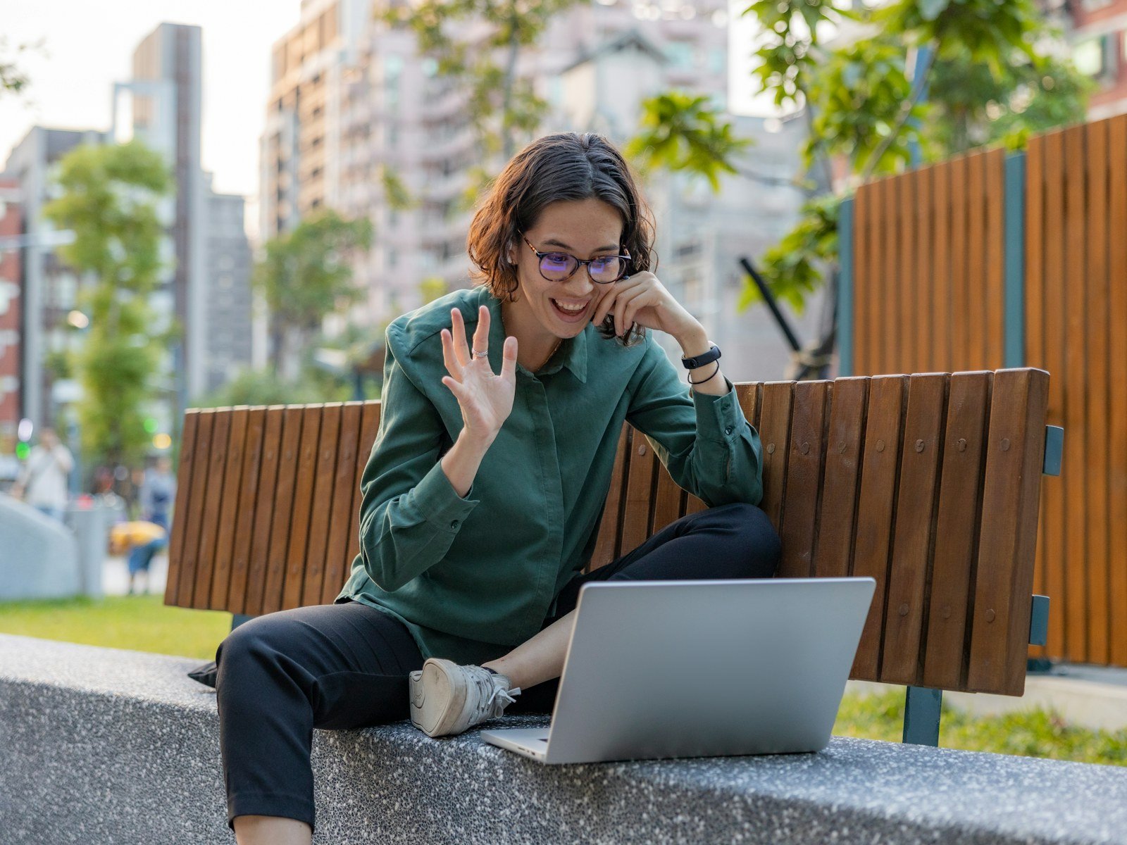 A woman waving hello on a video call while working outdoors on her laptop